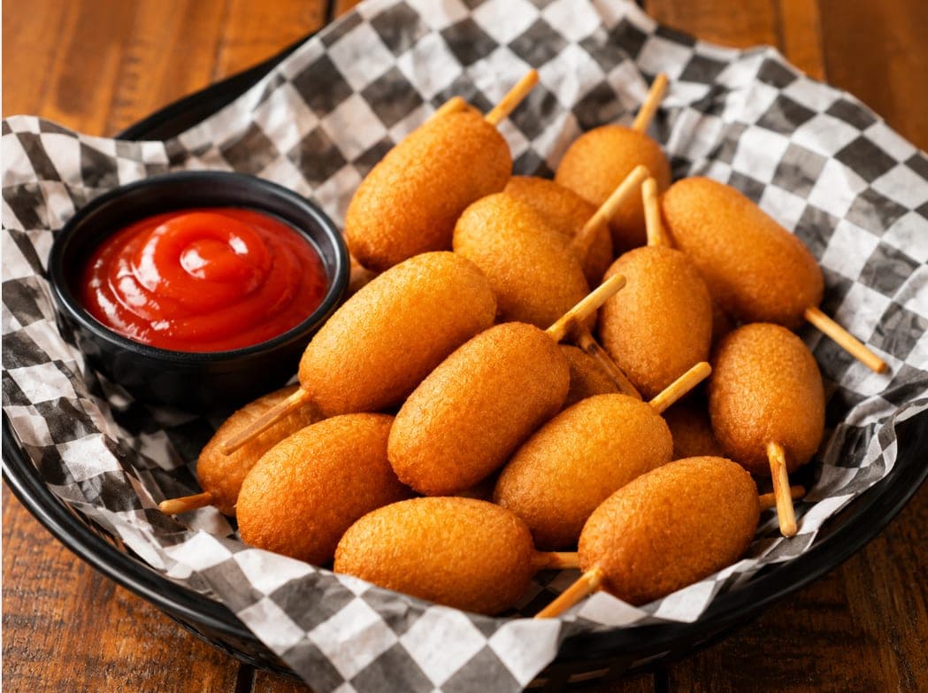 A basket of mini corn dogs on sticks with a side of ketchup.
