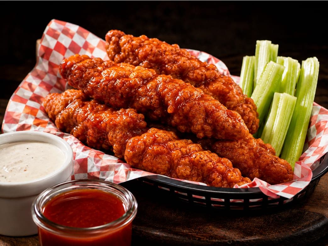 A basket of chicken tenders tossed in buffalo sauce, with celery and a side of ranch.