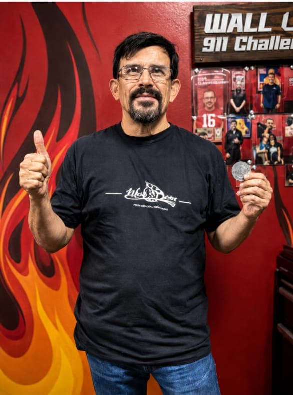A 911 Challenge winner giving a thumbs up with a Wicked Chicken coin beside the 911 Challenge wall.